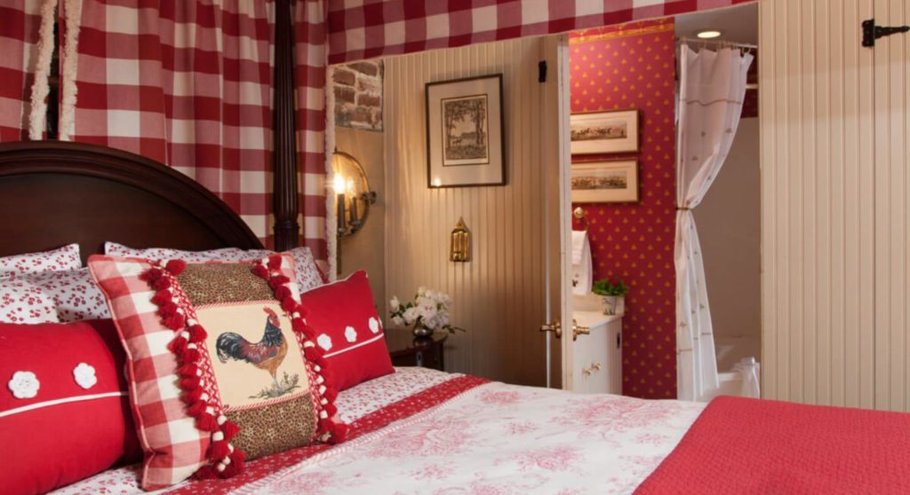 Bedroom with dark wooden four-poster canopy bed, red and white bedding, white-paneled walls, and view into bathroom