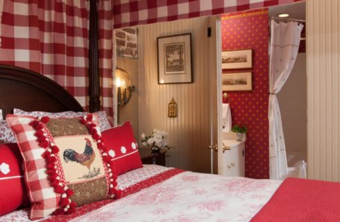 Bedroom with dark wooden four-poster canopy bed, red and white bedding, white-paneled walls, and view into bathroom