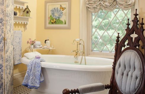 Bathroom with white soaker tub, yellow walls, ornate dark wooden chair, and window with a view to the outside