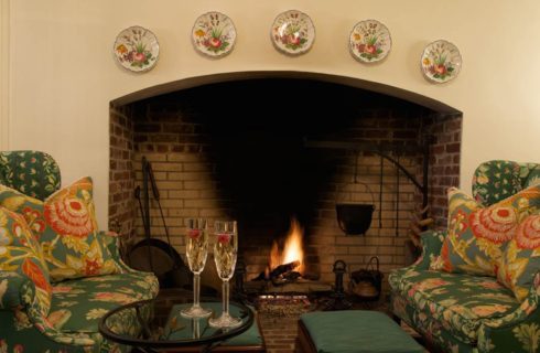 Close up view of red, pink, and green upholstered arm chairs and ottomans near large stone brick burning fireplace