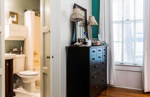 Bedroom with hardwood flooring, white and teal walls, dark wooden dresser, and view into bathroom with white tile flooring