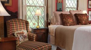 Large bedroom with dark wooden four-poster bed, white and tan bedding, antique upholstered love seat, and plaid upholstered armchair