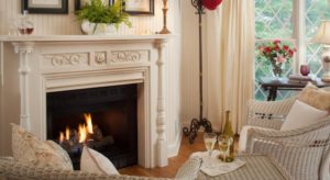 Close up view of white wicker chair and table near burning fireplace with white mantel and white wood-paneled walls