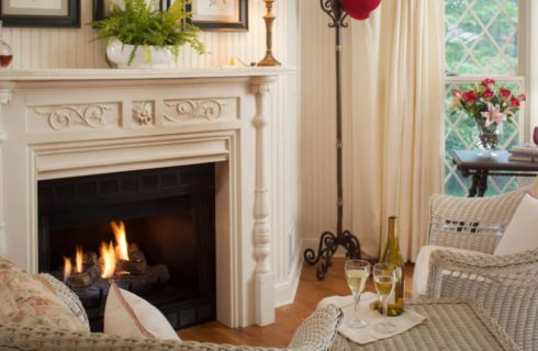 Close up view of white wicker chair and table near burning fireplace with white mantel and white wood-paneled walls