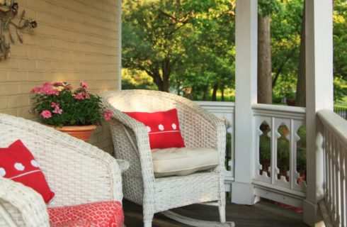 Front porch with white wicker rocking chairs and bright red pillows and green trees in the background