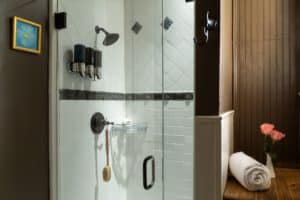 Shower with glass doors and mounted shampoo/conditioner/bodywash apparatus. White tile in the shower. Shelf on the right side of the image with a rolled white towel and small floral arrangement
