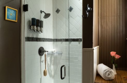 Shower with glass doors and mounted shampoo/conditioner/bodywash apparatus. White tile in the shower. Shelf on the right side of the image with a rolled white towel and small floral arrangement