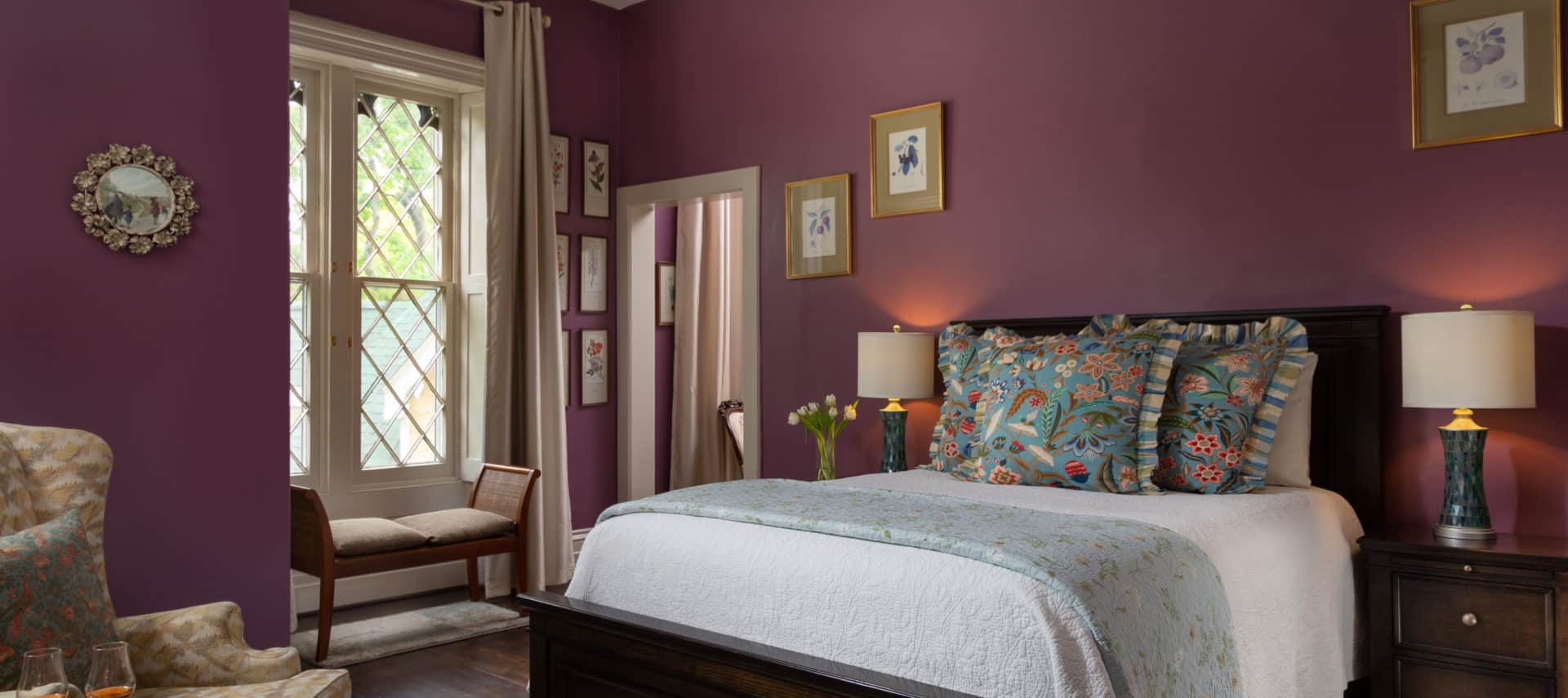 Spacious room with plum walls and a large gothic window with diagonal hatching. A queen sized bed on the right of the image with white bedspread and decorative pillows and on the left of the image is a chair and a side table with two glasses of bourbon. There is a bench under the window in the background.