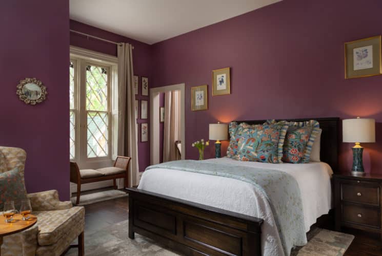 Spacious room with plum walls and a large gothic window with diagonal hatching. A queen sized bed on the right of the image with white bedspread and decorative pillows and on the left of the image is a chair and a side table with two glasses of bourbon. There is a bench under the window in the background.