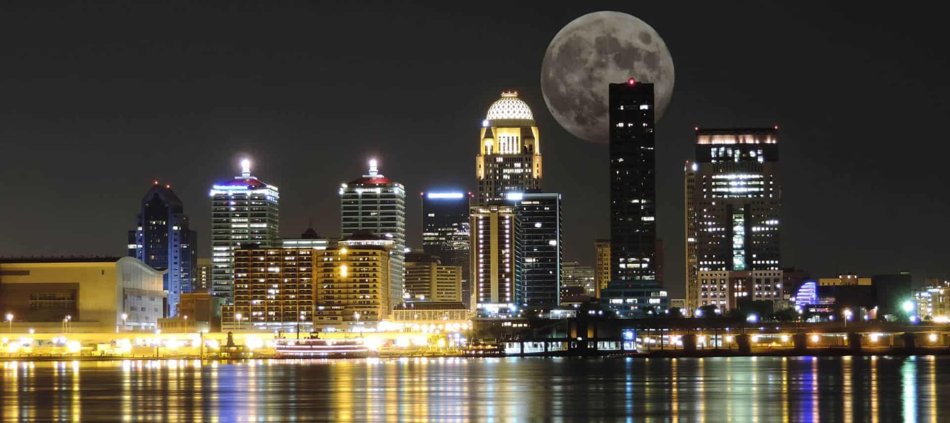 Skyline of city buildings lighted up at night near a body of water with a large moon in the background