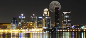 Skyline of city buildings lighted up at night near a body of water with a large moon in the background