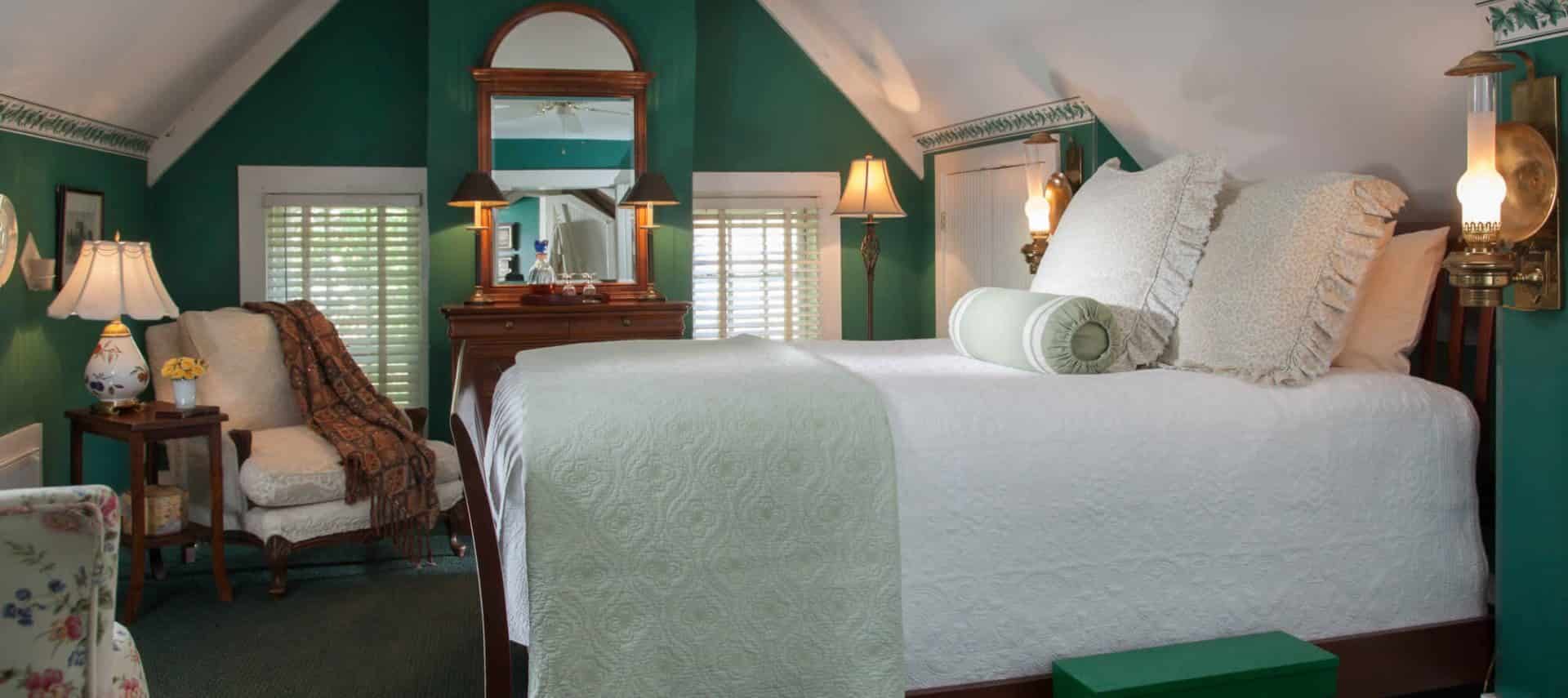 Large bedroom with hunter green walls and carpeting, dark wooden sleigh bed with white bedding, upholstered armchair, and wooden dresser