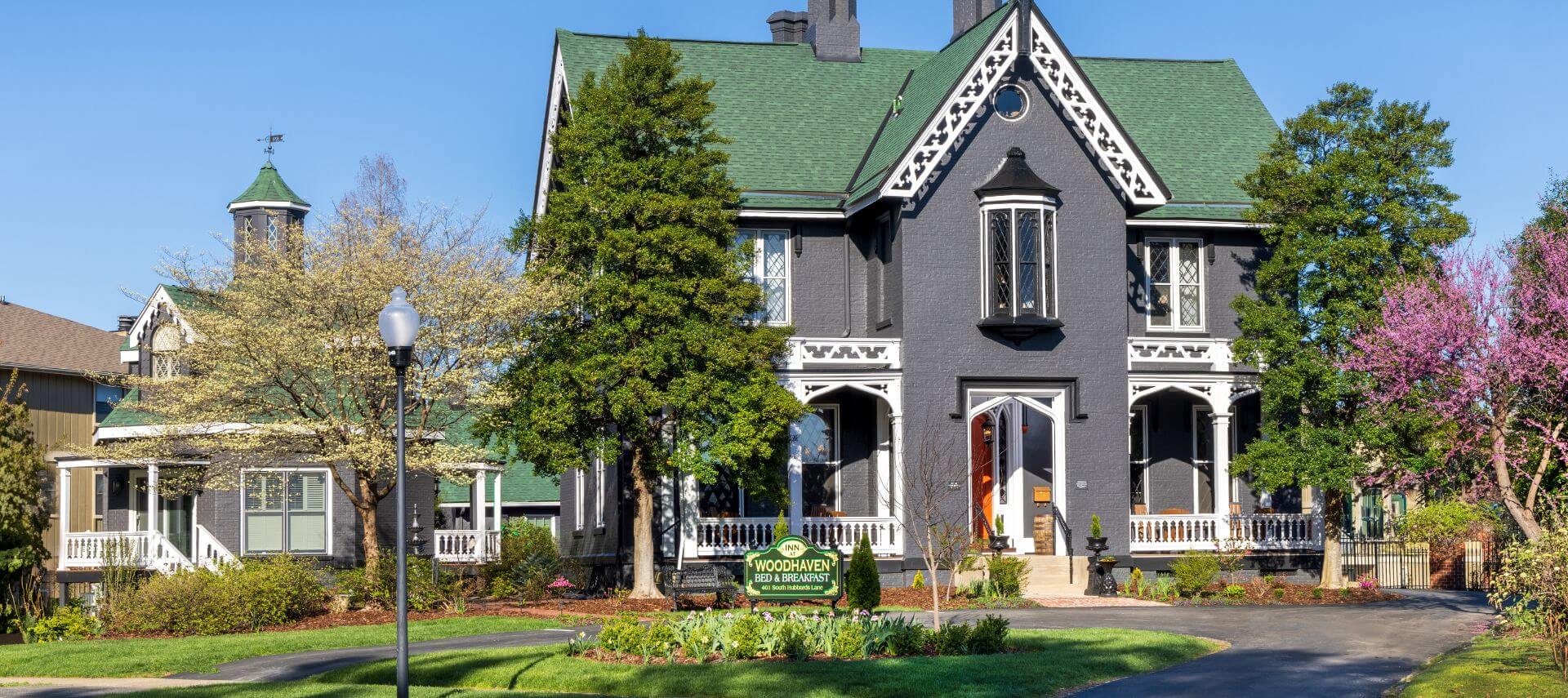 Exterior view of the property painted gray with white trim surrounded by green trees, shrubs, and grass, yellow and green sign in front