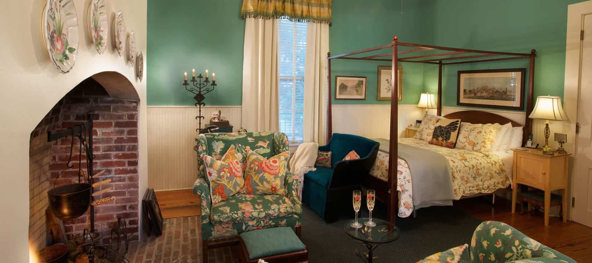 Large bedroom with white and green walls, hardwood flooring, dark wooden four-poster bed, upholstered chairs, and large red brick cooking fireplace