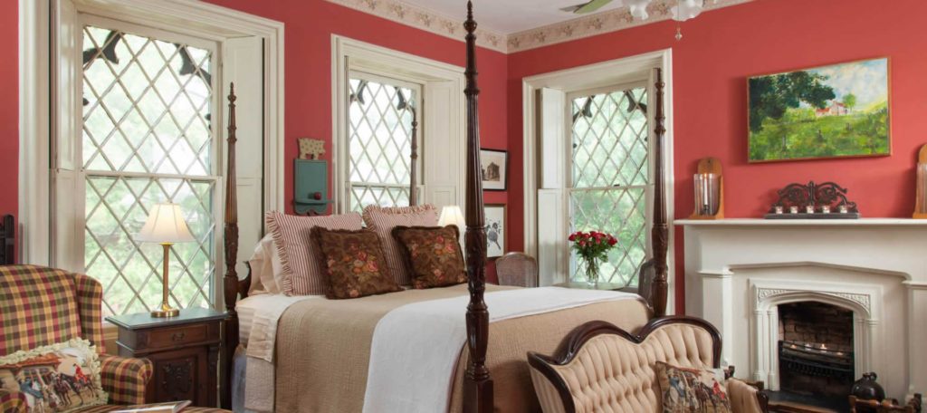 Large bedroom with dark wooden four-poster bed, white and tan bedding, antique upholstered love seat, plaid upholstered armchair, and fireplace