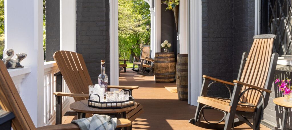 long view of multiple porches with wooden adirondack chairs and rockers at gray house, front porch with wooden stand holding refreshments.