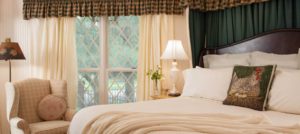 Bedroom with dark wooden headboard, white bedding, light tan upholstered armchair, and large window with view to the outside