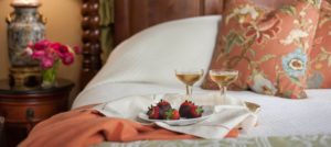 Close up view of white plate with chocolate covered strawberries and glasses filled with wine sitting on tray on bed with white bedding
