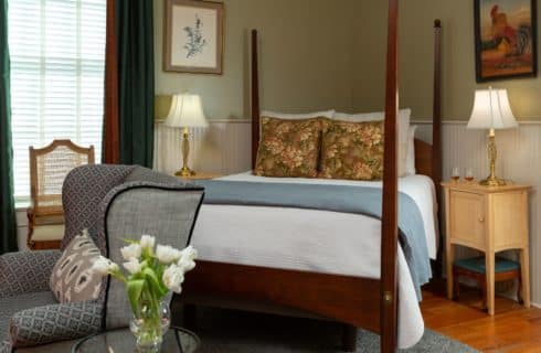 Foreground of side table next to a large gran chair with an arrangement of white flowers. Background of queen sized bed with decorative pillows