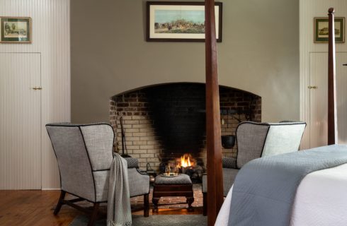 sitting area in front of a lit fireplace with two gray over-sized chairs and a throw tossed over the arm of one chair. An ottoman sits between the chairs with two glasses of bourbon on it.