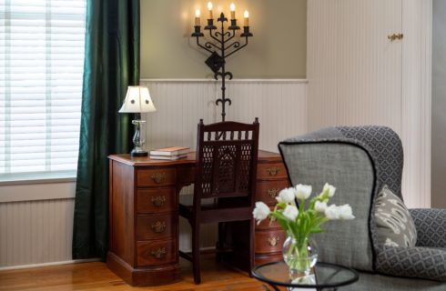 Wood desk with a dark wood chair. White lamp on the desk with a crystal vase of white flowers in front.