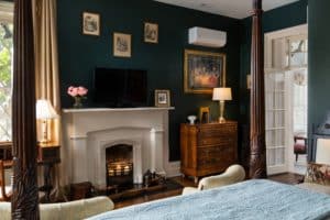 View from the queen four-post bed looking at the fireplace. Electric candles placed in the fireplace. A dresser sits to the right of the fireplace and period victorian pictures are on the wall above the mantle.