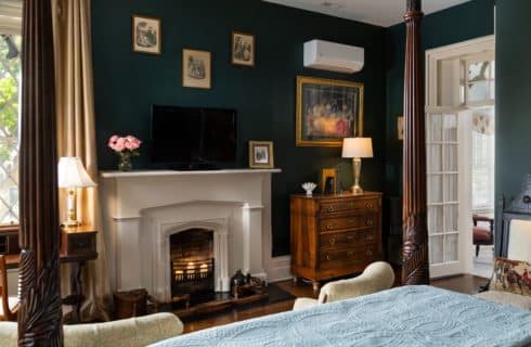 View from the queen four-post bed looking at the fireplace. Electric candles placed in the fireplace. A dresser sits to the right of the fireplace and period victorian pictures are on the wall above the mantle.