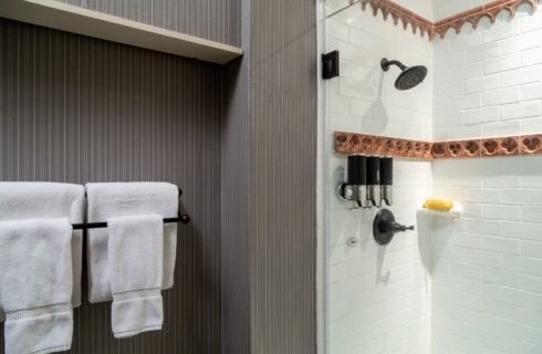 Picture of shower area with gray pinstripe wallpaper and tall steam shower with white towel and mounted shampoo/conditioner/bodywash apparatus. Glass shower doors and white towels hanging on a towel bar outside the shower area.