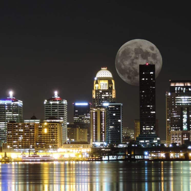Skyline of city buildings lighted up at night near a body of water with a large moon in the background