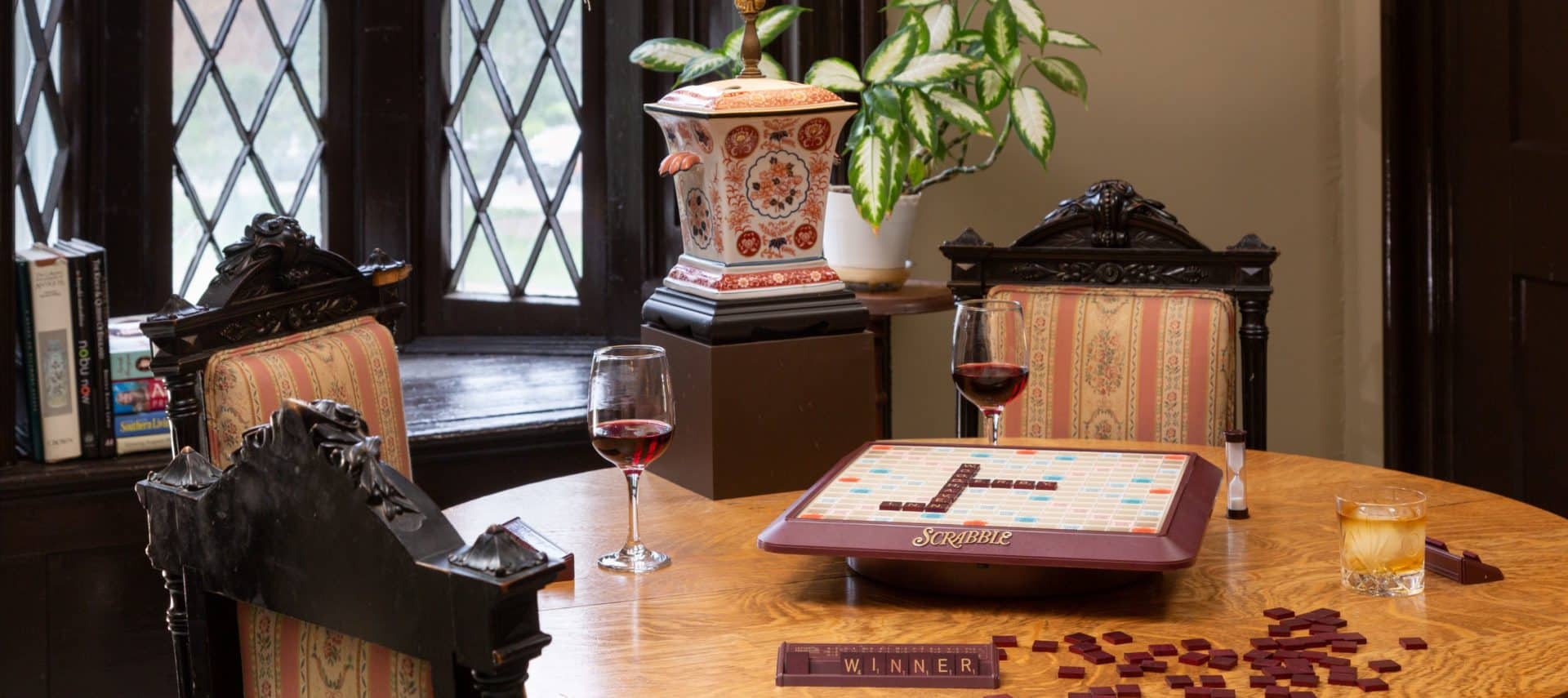 Game table with Scrabble tiles spelling winner. A glass of wine and a glass of bourbon are on the table with a gothic bay window behind the table.
