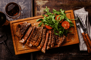 medium rare steak sliced into strips on a wooden cutting board with tomatoes and arugula.