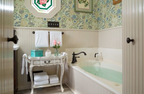 view of bathroom with large jetted soaking tub full of water, plush white towels and an arrangement of pink roses next to the bathtub. Walls are covered in pale green floral wallpaper