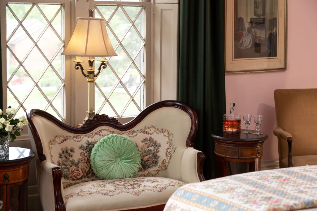 The foot of the bed is on the bottom right of the image and there is a victorian setee in front of two gothic style windows and a decorative lamp behind the setee.