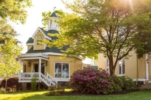 Daytime view of the front of the Rose Cottage. Octagonal structure painted yellow with a green roof and white gingerbreading. Surrounded by lush green landscaping.