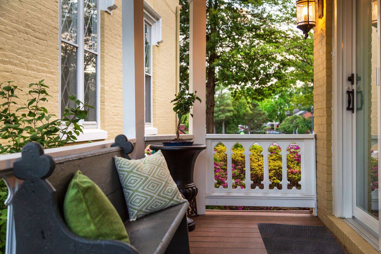 A patio area with a gray bench and two decorative pillows.  A white rail in the background with pink colorful flowers visible through the rails. 