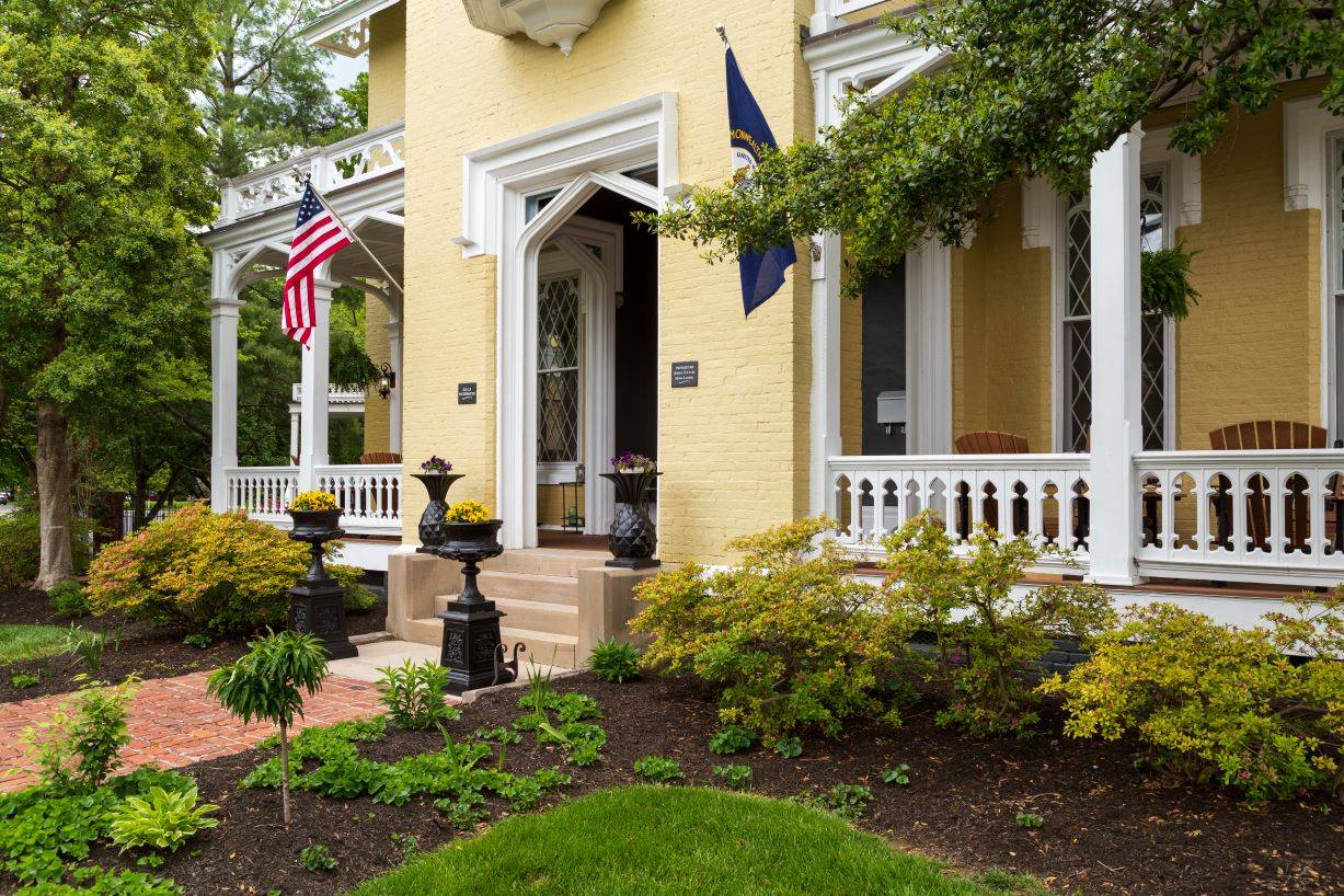 View of front of house.  Yellow and white with two flags and black plant boxes and lush green landscaping. 
