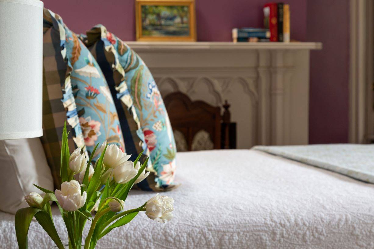 Side of a bed with white coverlet and light blue pillows.  An arrangement of white flowers is on the nightstand. 