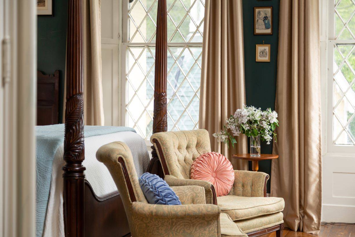 View of the end of a four-post bed with two chairs and decorative colorful pillows on the chairs.  A large gothic window is in the background. 