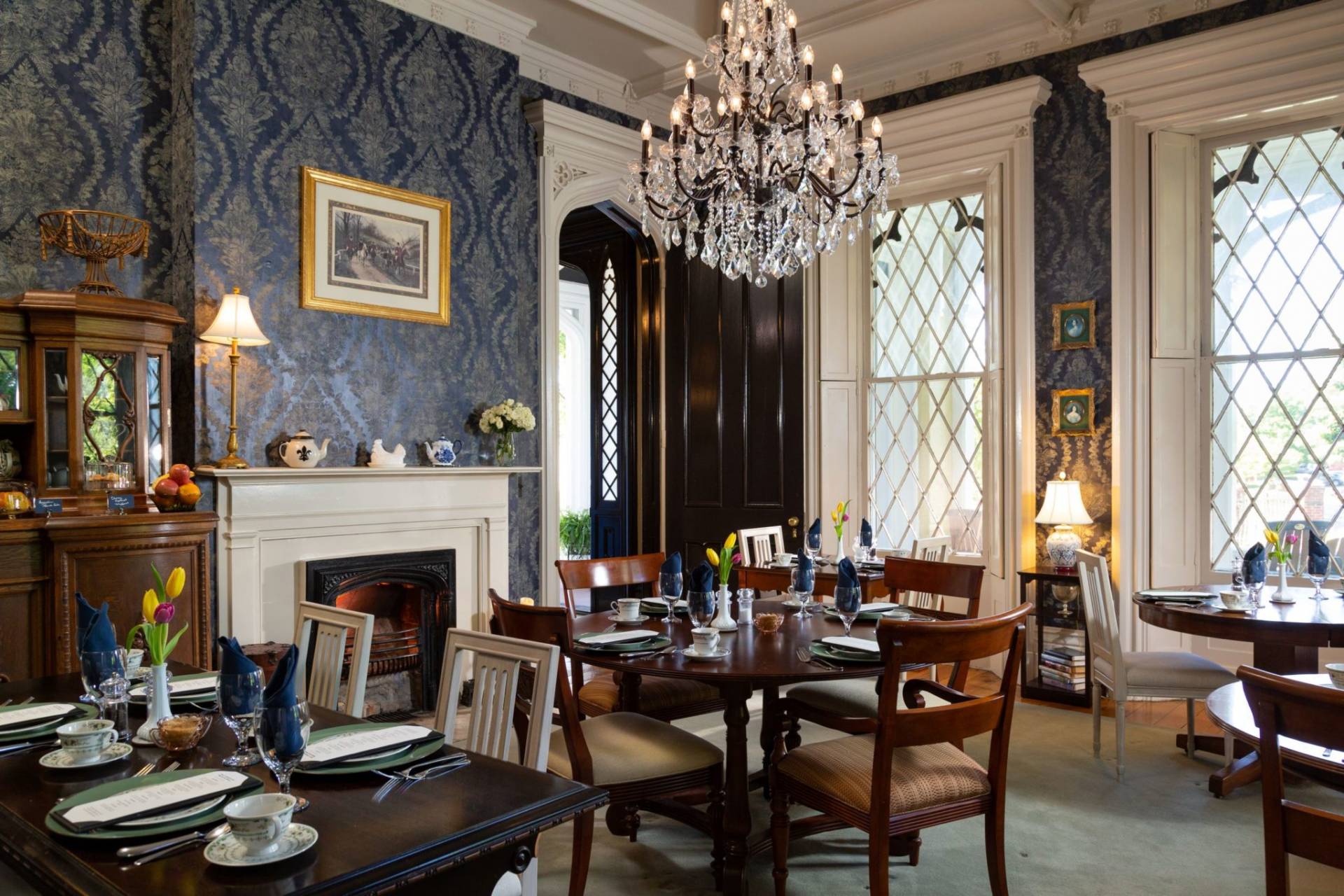 Dining room with multiple wood tables set for breakfast.  Navy historic wallpaper and a fireplace.  A large crystal chandelier in the center of the picture.