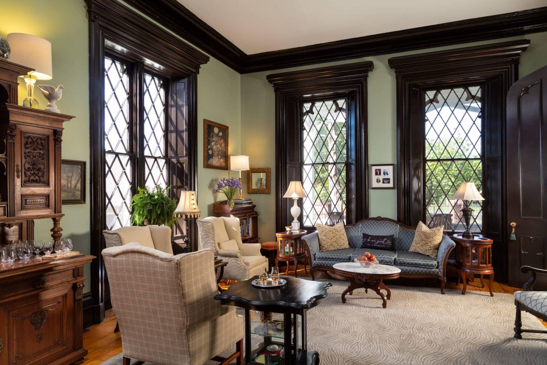 Living room with pale green walls and four large gothic style windows.  Multiple seating and coffee tables and two glasses of bourbon. 