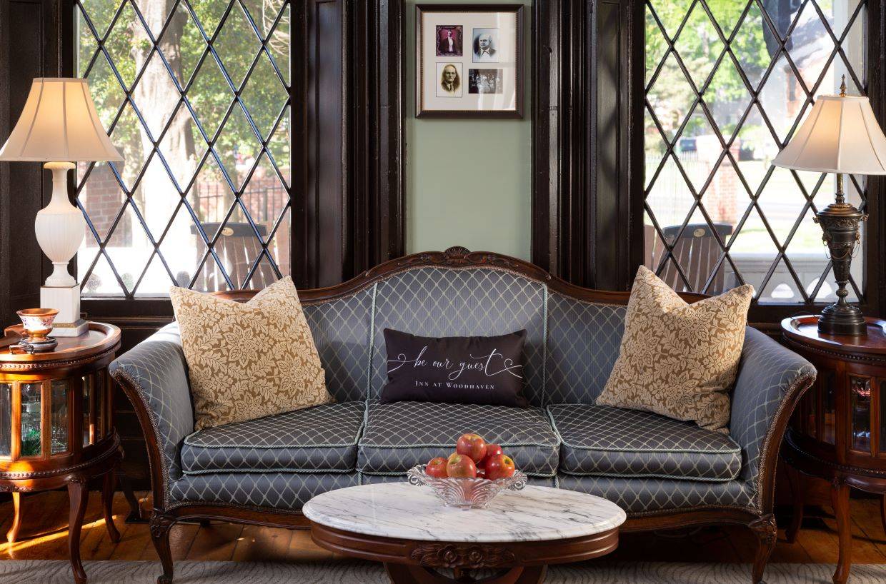 A blue sofa with decorative pillows sits in front of two very large gothic style windows and a marble coffee table sits in front of the sofa with a bowl of red apples. 