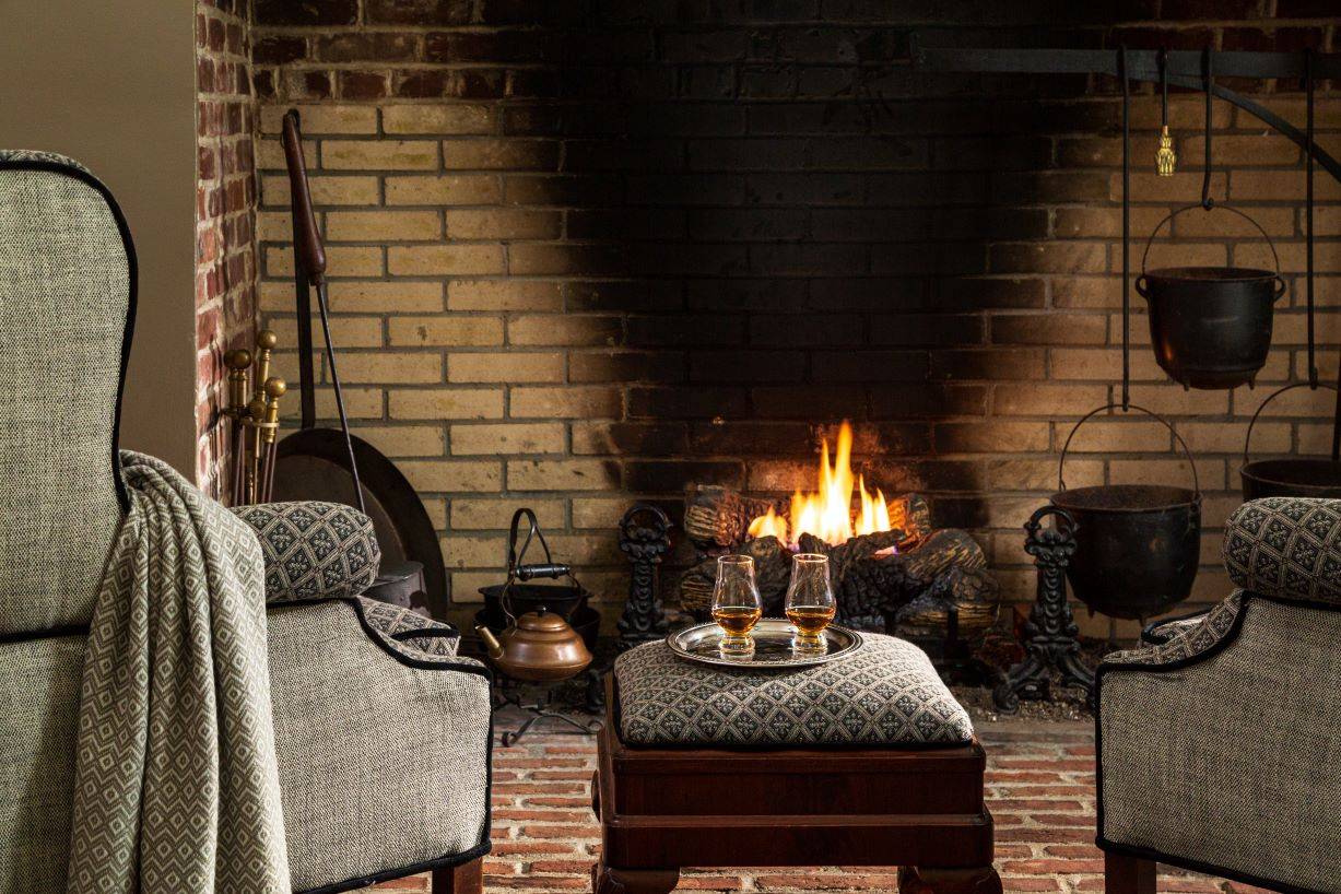 A gray chair in front of a fireplace with an ottoman and two glasses of bourbon on  a tray in front of the fire. 