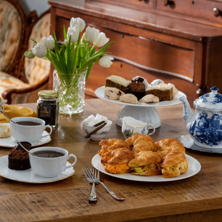 Close up view of tea set with croissant sandwiches, pastries, a teapot and two cups of tea and a floral arrangement of white daffodils.