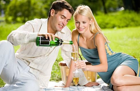 Man with white sweater pouring Champagne into glass held by woman with blue dress on picnic blanket