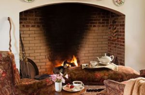 Close up view of red, pink, and brown upholstered arm chairs and ottoman near large stone brick burning fireplace