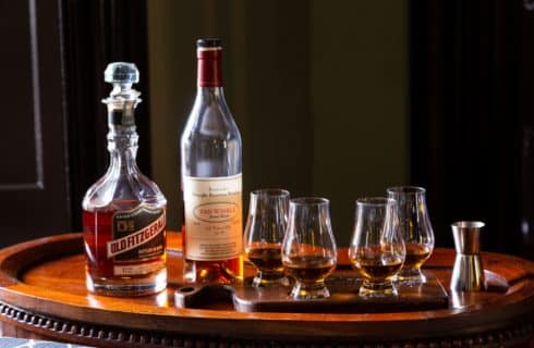 four tasting glasses containing bourbon on a wooden flight board with two bottles of bourbon
