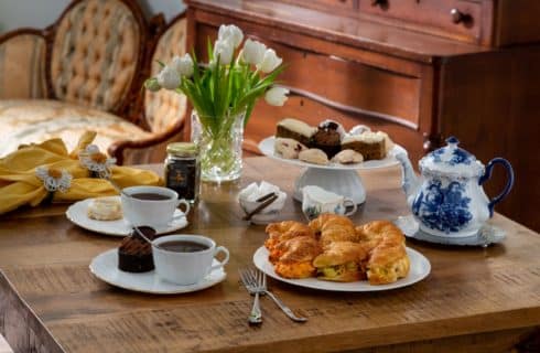 set table with ornate teapot, vase of flowers, croissant sandwiches, tea and cookie selection