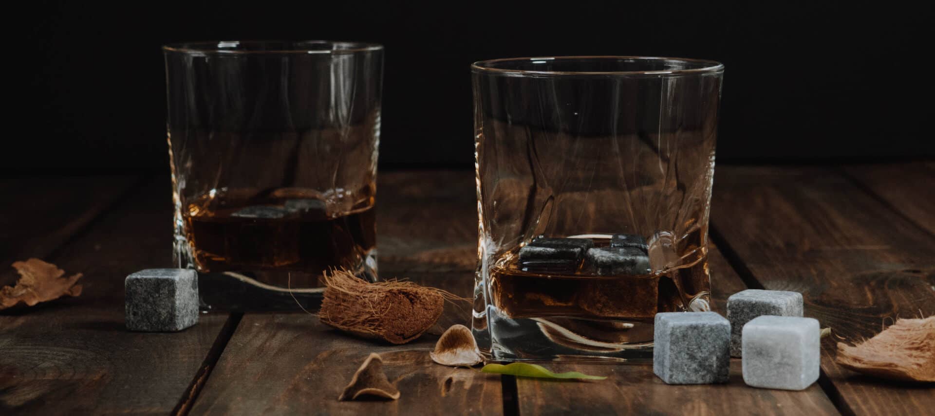 Two rocks glasses with golden bourbon whiskey on a wooden table covered in natural materials and four grey whiskey stones.