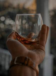 Caucasian hand with a leather wristwatch swirling a stemless round glass with amber-colored bourbon to look at the whiskey’s legs.