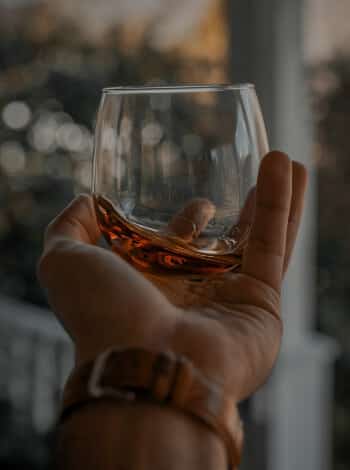 Caucasian hand with a leather wristwatch swirling a stemless round glass with amber-colored bourbon to look at the whiskey’s legs teaching people how to drink bourbon 